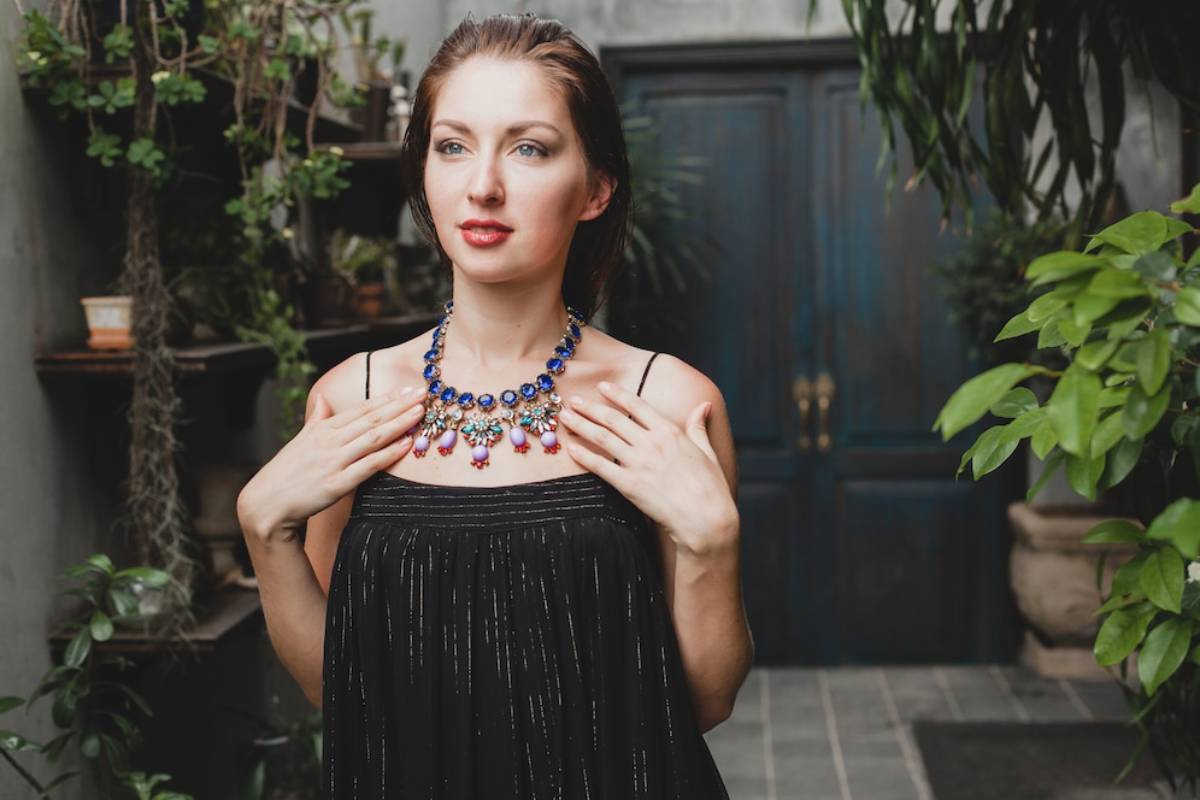 Woman in a black dress touching a colorful necklace, standing in a garden with plants.