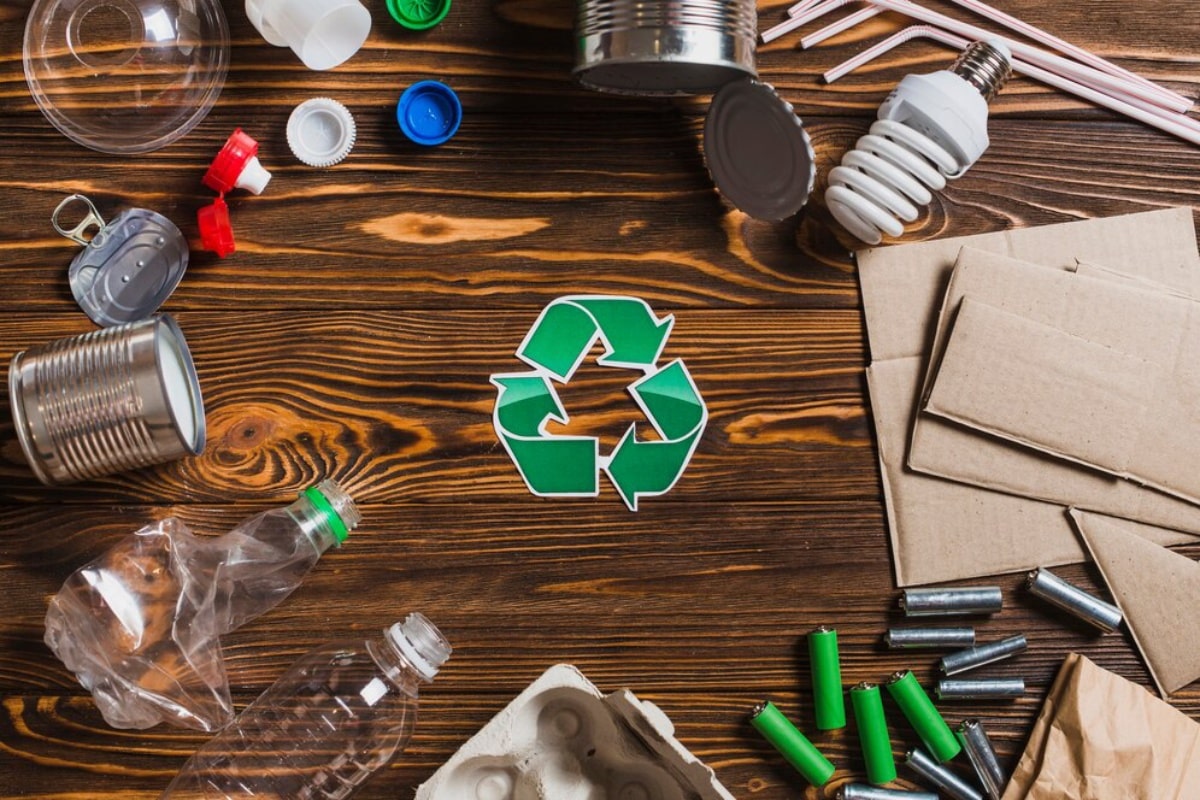 Assorted recyclables such as plastics, metals, and paper arranged around a recycling symbol on a wooden surface.