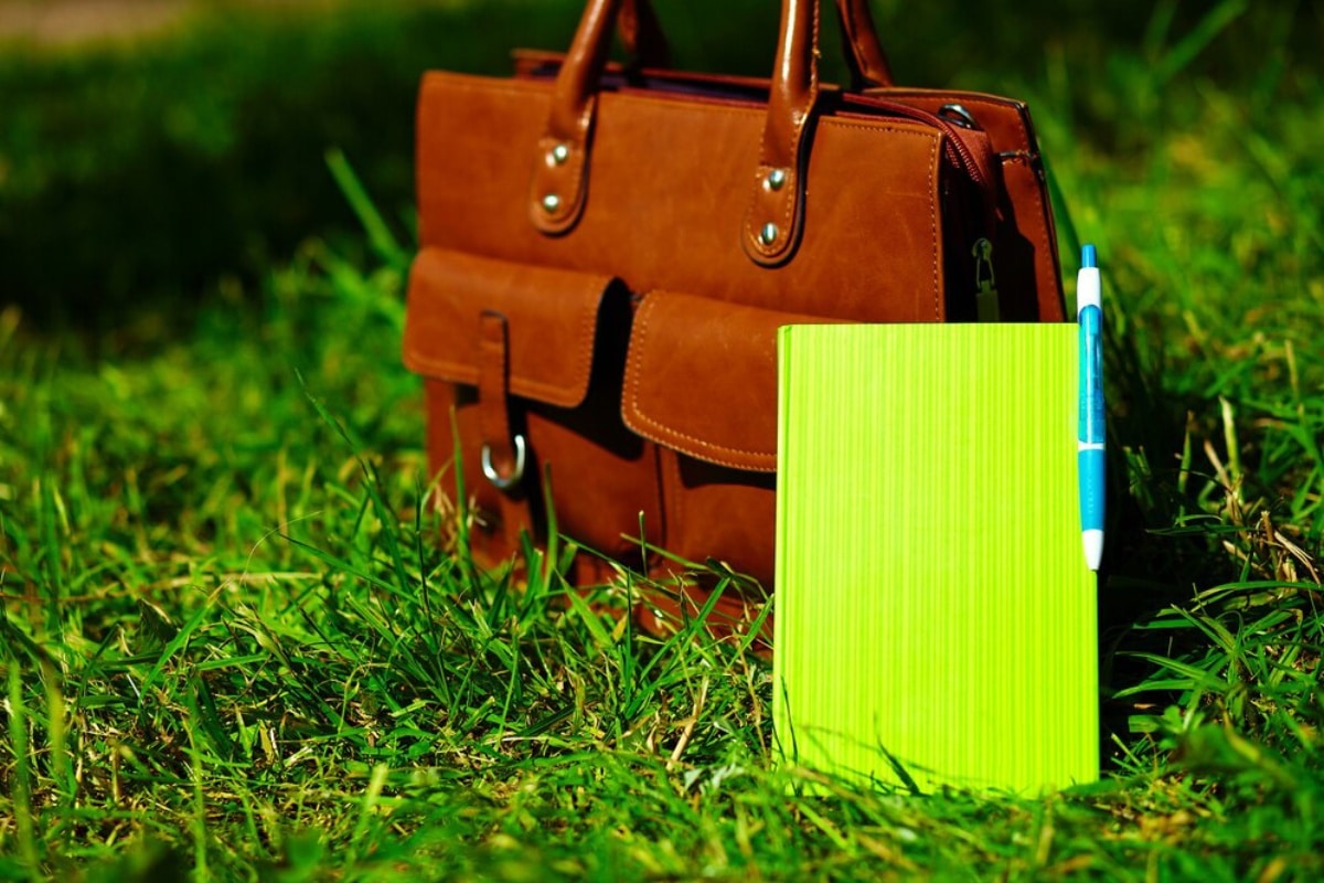 A brown leather bag and a neon green notebook with a blue pen on grass.
