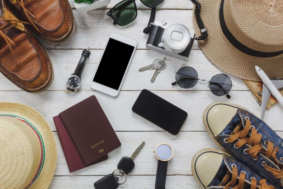 Travel essentials laid out on a wooden surface, including shoes, sunglasses, camera, smartphone, passport, and hat.