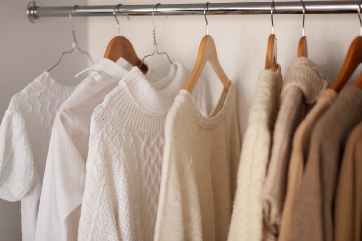 Neutral-toned sweaters and shirts hanging on wooden hangers on a metal rail.
