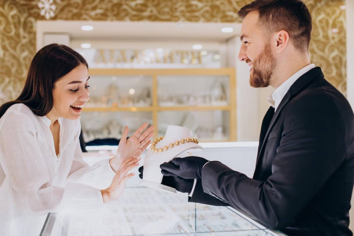 A man in a suit presents a jewelry box to an excited woman in a store.