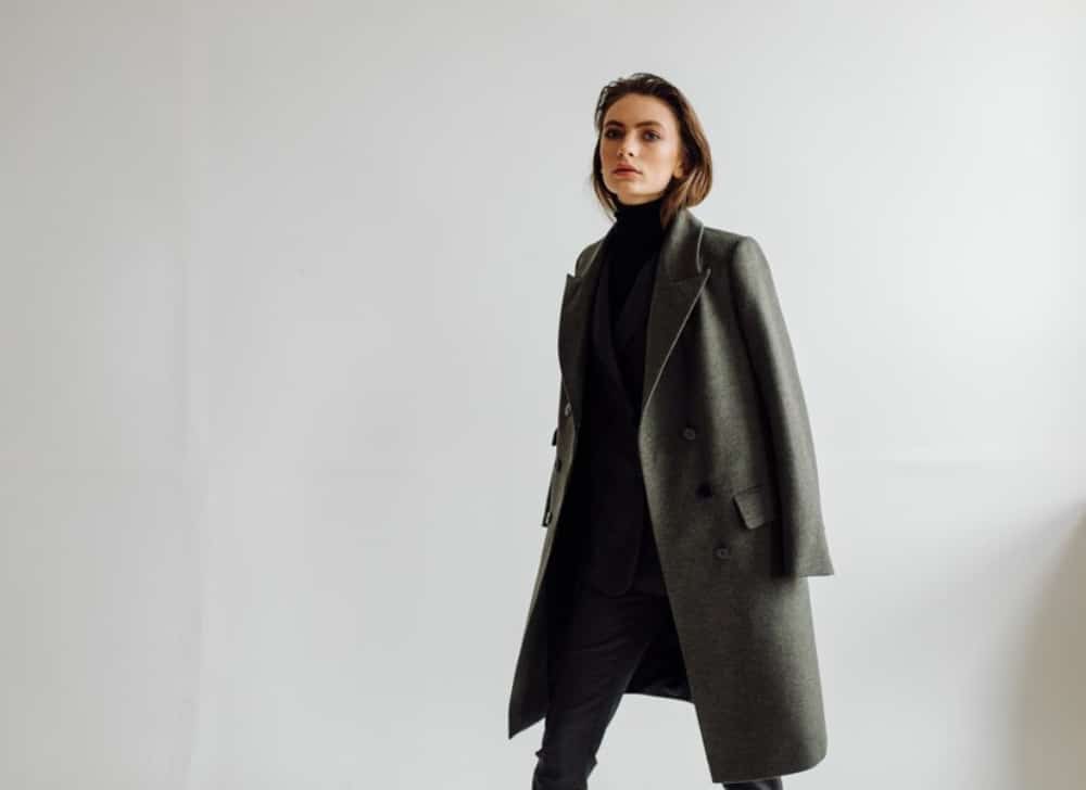 Woman in stylish grey coat and black turtleneck, posing in a minimalist white setting.