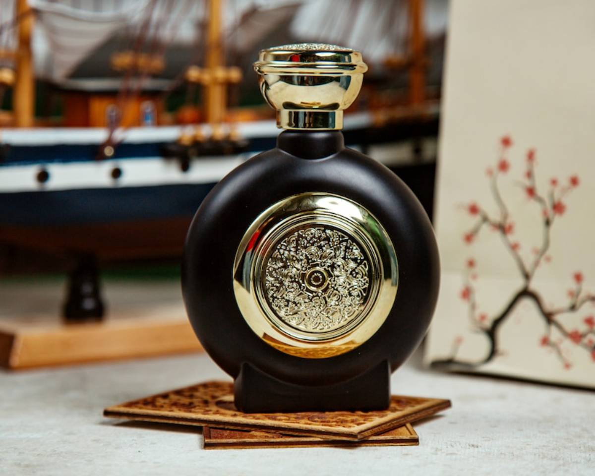 Ornate black and gold perfume bottle on a wooden stand, with a ship model and floral painting in the background.