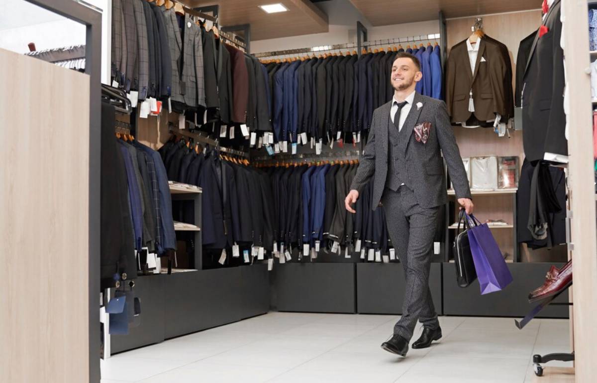 Man in a grey suit shopping happily in a menswear store, browsing suits and holding a purple shopping bag.