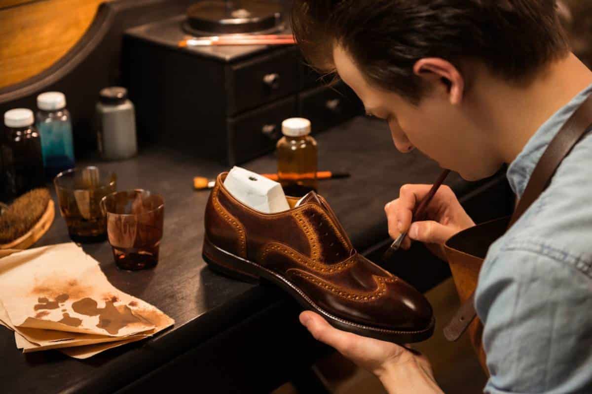A craftsman meticulously paints details on a brown leather shoe in a workshop.
