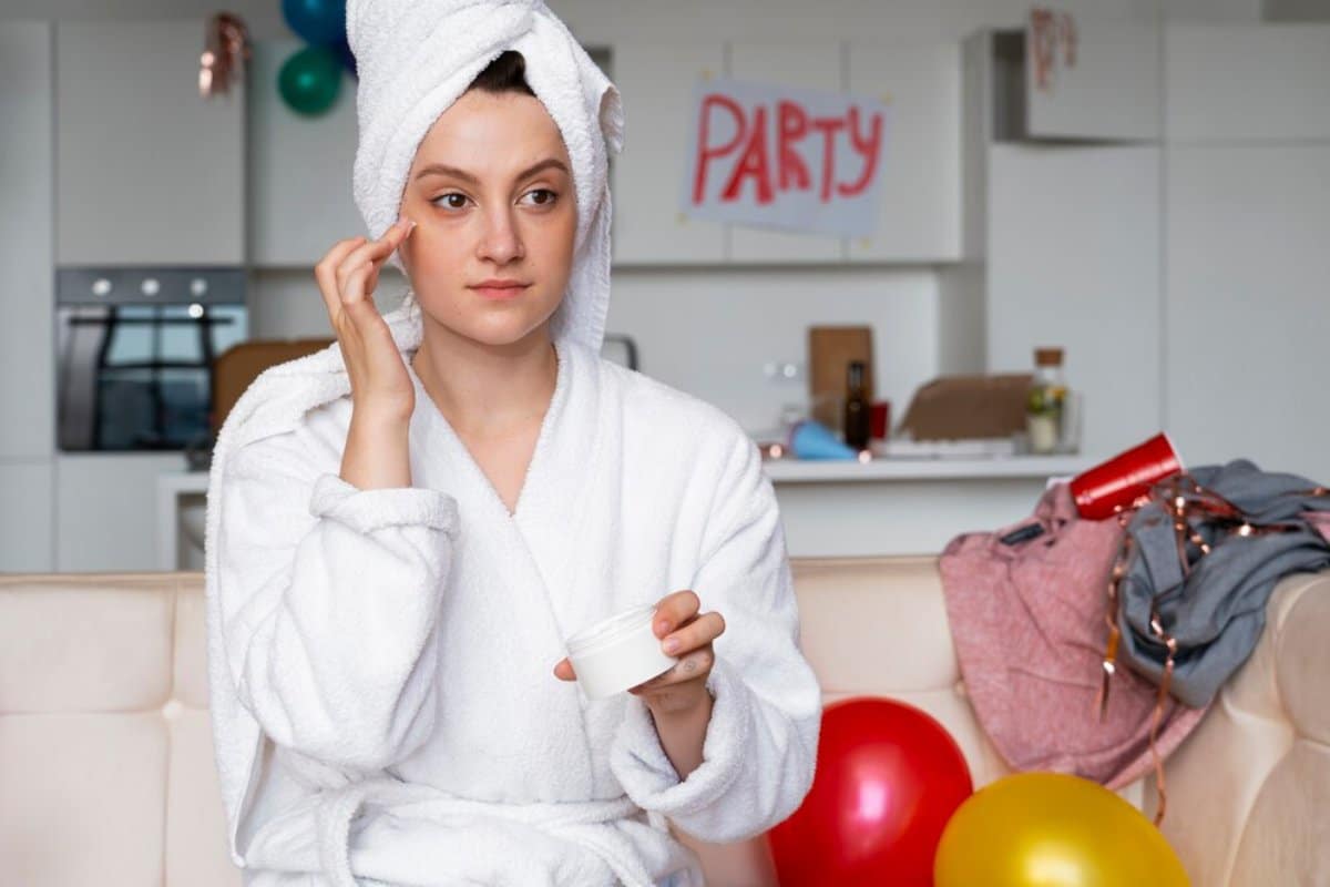Woman in white robe and towel on head applying cream on face in a living room decorated with "PARTY" sign and balloons.