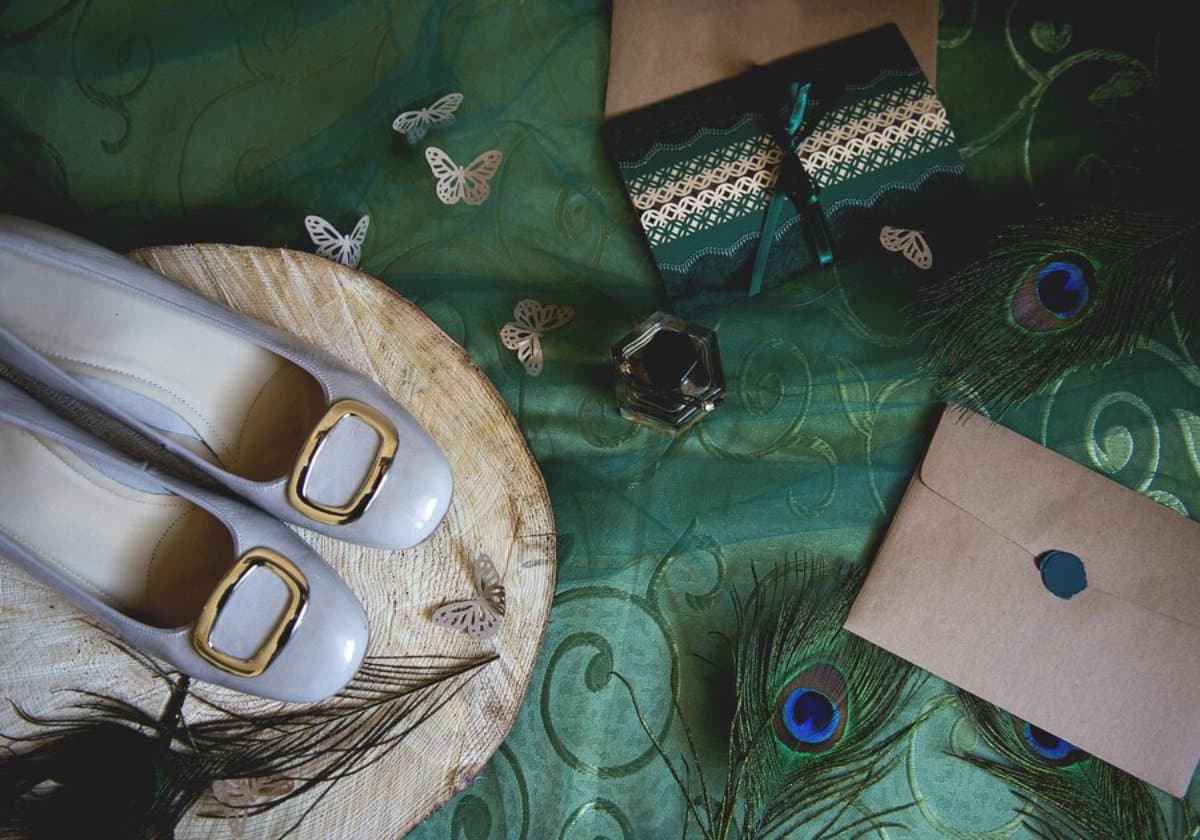 Elegant women's shoes, perfume bottle, and gifts on a green patterned fabric with butterfly accents and peacock feathers.