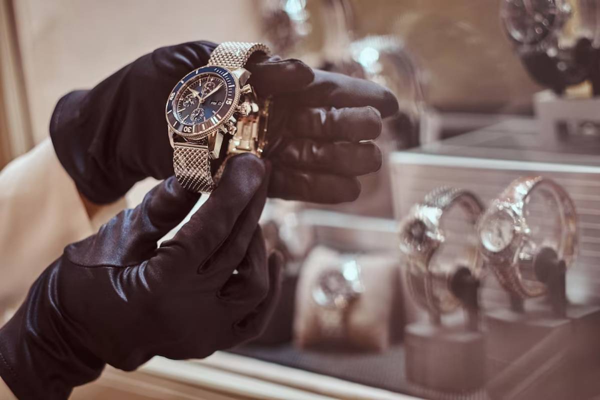 Hands in black gloves holding a luxurious wristwatch in front of a display case with blurred watches in the background.