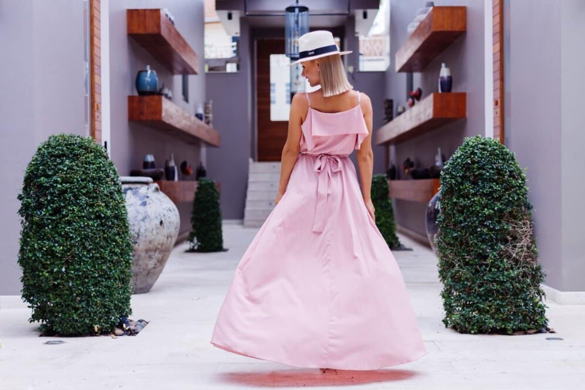 Woman in a pink dress and white hat stands in a stylish corridor lined with green topiaries and decorative shelves.