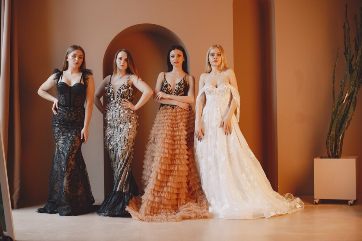 Four women in elegant evening gowns posing in a room with warm-toned walls and a tall cactus plant.
