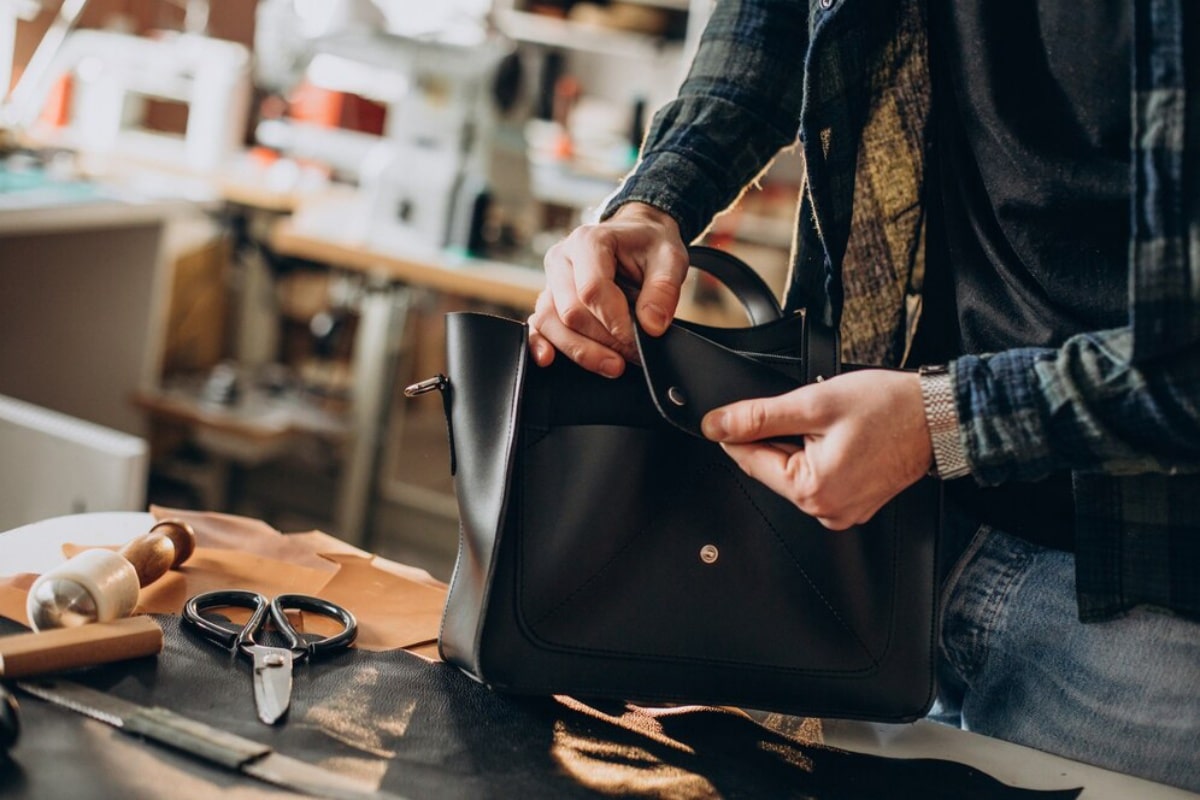 A craftsman working on a black leather handbag in a workshop, surrounded by tools.