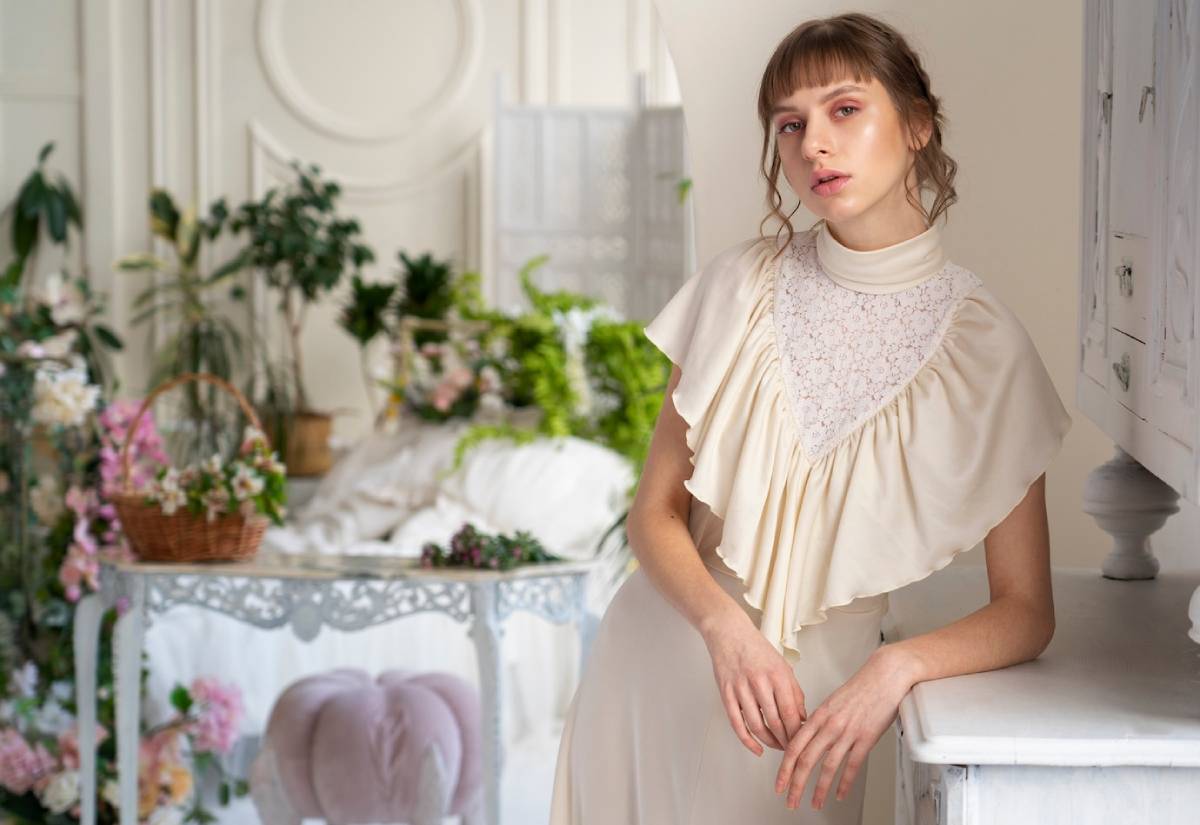 A young woman in a vintage cream blouse stands near a table in a floral-decorated room.