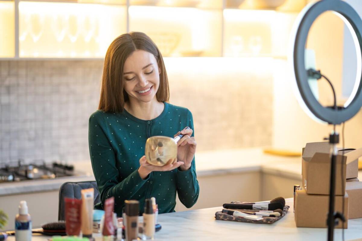 Woman reviews makeup products while recording a video in a bright kitchen setting.