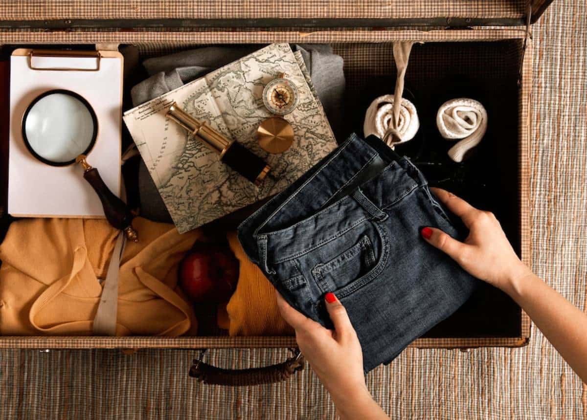 Hands packing a suitcase with a denim jean, map, magnifying glass, compass, and clothing for travel.