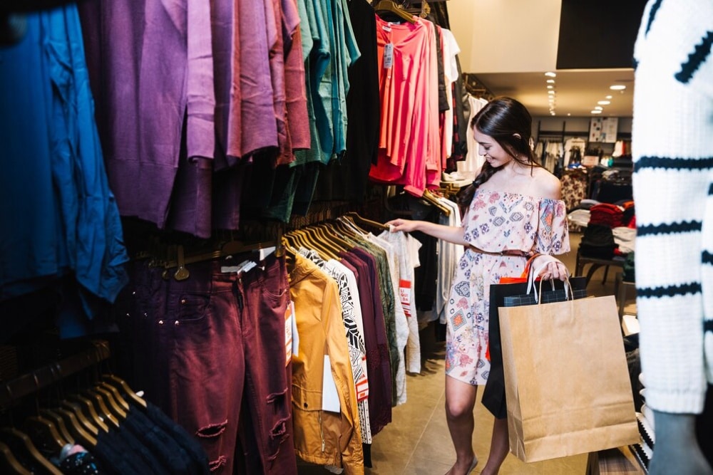 Woman shopping, browsing through a rack of colorful clothing in a retail store, holding a shopping bag.