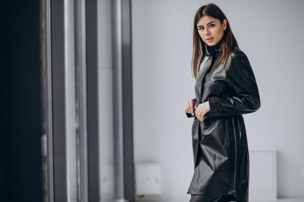 Woman in a sleek black leather coat, standing confidently in a modern, minimalist interior.